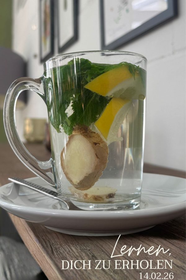 Das Bild zeigt eine Glastasse mit Ingwerscheiben und Minze auf einem Holzregal, das an einer Wand befestigt ist. Bilder sind im Hintergrund erkennbar.